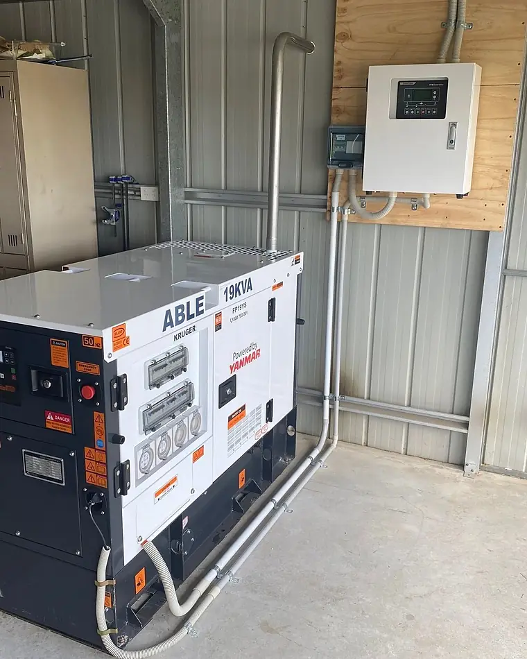 A diesel standby generator in a shed with its changeover board on the wall